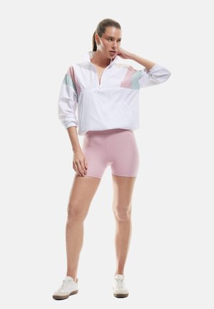 Rompevientos blanco con acentos en tonos pastel de rosa y menta, con un cuello de media cremallera. Unos pantalones cortos ajustados rosas claros y zapatillas blancas completan el conjunto.