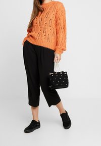 Dzianinowy pomarańczowy sweter o luźnym kroju i otwartym wzorze, zestawiony z czarnymi spodniami o długości 3/4 oraz czarnymi sneakersami, trzymający małą, pikowaną czarną torebkę.
