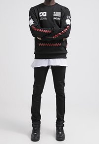 Maglione nero con grafiche stampate e dettagli a zigzag rossi, indossato sopra una maglietta bianca. Abbinato a jeans neri e sneakers nere.