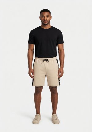 Homme debout, faisant face, portant un t-shirt noir, un short beige avec des panneaux latéraux noirs et des baskets beiges sur un fond blanc.