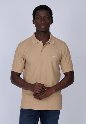 Polo en coton beige avec un col, deux boutons, des manches courtes et un petit logo brodé sur la poitrine. Porté avec un jean noir.