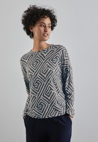 Sweter z długim rękawem w beżowym i granatowym kolorze z geometrycznym wzorem. Miękka tekstura, okrągły dekolt i luźny krój. Połączony z ciemnymi spodniami.