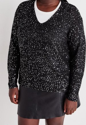 Personne portant un pull noir à paillettes avec col en V sur une chemise blanche, associé à une jupe en cuir noire, debout les mains détendues.