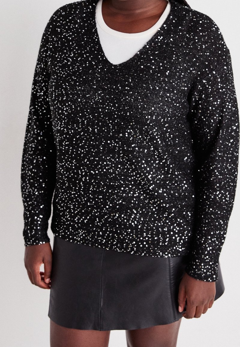 Personne portant un pull noir à paillettes avec col en V sur une chemise blanche, associé à une jupe en cuir noire, debout les mains détendues.