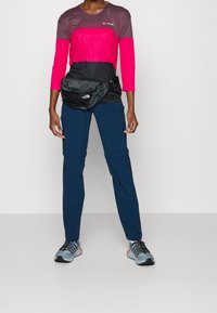 Prestanda idrottskläder med en färgad långärmad topp i rosa och svart, matchad med marinblå byxor och en svart midjeväska.