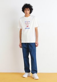 T-shirt branca com o texto "A Escolha Inteligente" e o logótipo da New Balance, combinada com calças de ganga azul-escuro e ténis brancos.
