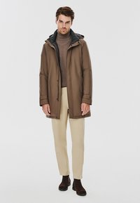 Brązowy ocieplany park z kapturem, nałożony na beżowy sweter. Jasne spodnie i brązowe botki za kostkę dopełniają zestaw, stojąc na szarym tle.