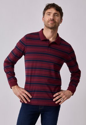 Hombre con barba que lleva una camiseta polo de manga larga a rayas burdeos y azul marino con las manos en las caderas, sobre un fondo liso.