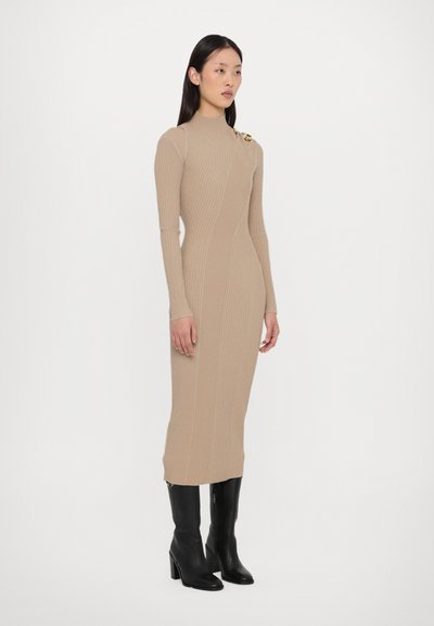 Beige ribbed knit dress with long sleeves, high neckline, side cutouts, and a decorative accent on the shoulder; paired with black heeled boots.