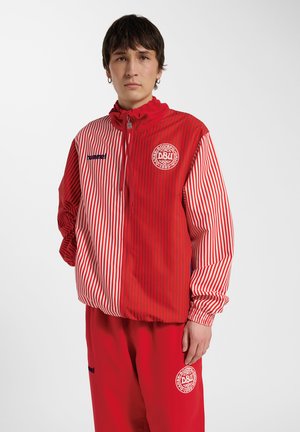 Jonge man met een rood-wit gestreept sportjack en rode broek met DBU-logo's, staand tegen een effen witte achtergrond.