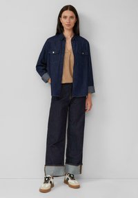 Donkerblauw denim shirt met twee borstzakken en opgerolde mouwen; gecombineerd met donkere, wijde spijkerbroek en meerkleurige sneakers.