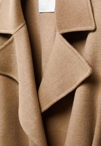 Gros plan sur le col d’un manteau en laine camel avec de larges revers, mettant en valeur le tissu texturé et l’étiquette en laine.