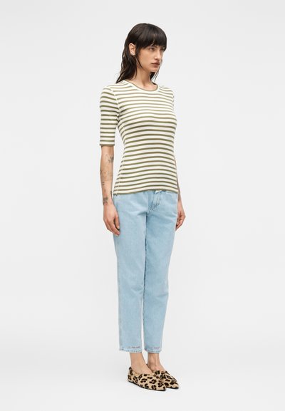 Γυναίκα με σκούρα μαλλιά που φοράει πράσινη και λευκή ριγέ κοντομάνικη μπλούζα, ανοιχτό μπλε τζιν και φλατ παπούτσια με λεοπάρ σχέδιο, στέκεται μπροστά σε λευκό φόντο.