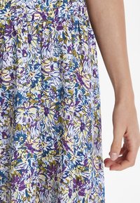 Abito floreale con design plissettato in bianco, con motivi floreali in viola, blu e giallo. Tessuto dalla superficie liscia. Mano visibile.