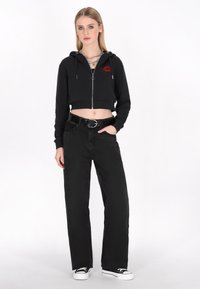 Sudadera negra corta con cremallera frontal y gráfico de labios rojos, combinada con jeans negros de pierna ancha de cintura alta y zapatillas negras.