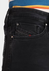 Vaqueros de mezclilla negra con textura desgastada, que presentan un bolsillo frontal y una pequeña etiqueta de marca, complementados con herrajes plateados en la cintura.