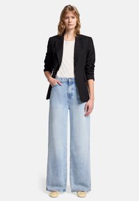 Blazer nero, t-shirt bianca e jeans in denim blu a gamba larga. Il denim presenta una finitura sbiadita. Scarpe di colore chiaro. Mani nelle tasche.