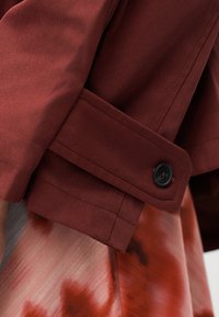 Manche d'un manteau rouge foncé avec un détail de poignet boutonné sur un fond de tissu à motif tie-dye rouge et blanc.