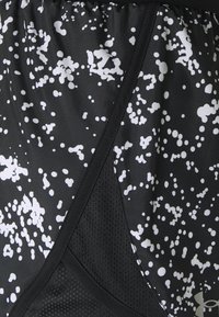 Gros plan sur un tissu athlétique noir avec motif blanc éclaboussé et détail en mesh, avec le logo Under Armour dans le coin inférieur droit.