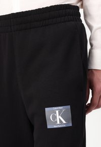 Zwarte sweatpants met een elastische tailleband, met een lichtblauw rechthoekig label dat "Calvin Klein" en "New York City" zegt.