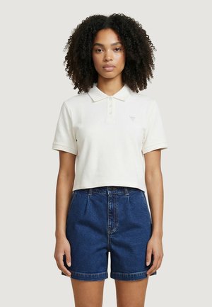 Joven con cabello rizado que lleva una camisa polo blanca de manga corta y corta, y pantalones cortos de mezclilla azul de cintura alta, posando contra un fondo liso.