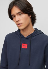 Mørkeblå hoodie laget av bomull med en rød logopatch merket "HUGO." Har snor i hetten og ribbestrikkede mansjetter. Glatt stofftekstur.