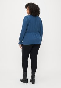 Blouse bleue texturée à manches longues et taille froncée, associée à des leggings noirs avec des détails sur les côtés et des bottines.