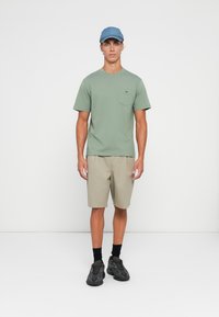 Een jonge man staat met een lichtgroen overhemd met korte mouwen en een zak, beige shorts, een blauwe pet, zwarte sokken en donkere sport schoenen voor een witte achtergrond.