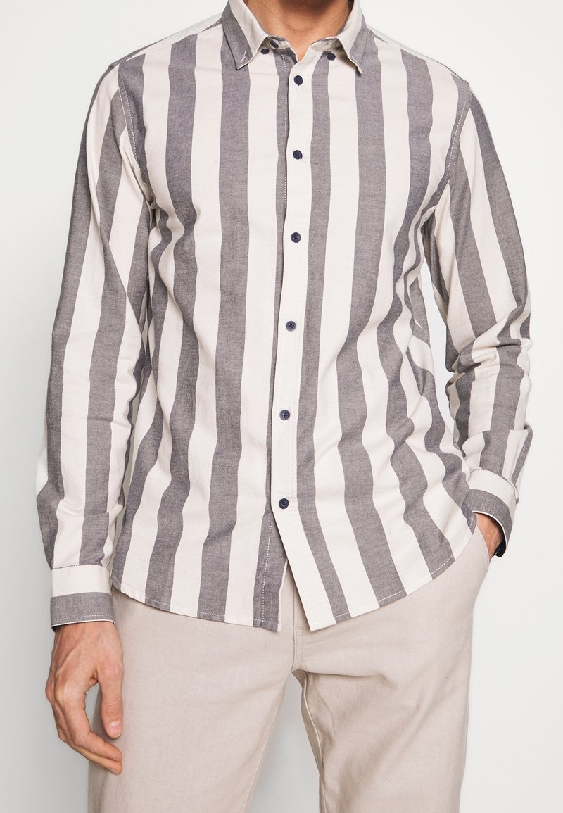 Homme portant une chemise à manches longues boutonnée avec de larges rayures verticales grises et blanches, associée à un pantalon clair.