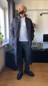Dolga črna puhovka z zadrgo, oblečena čez belo majico in črne hlače. Ima velike žepe in sproščen kroj. Črne čevlje zaključujejo videz.