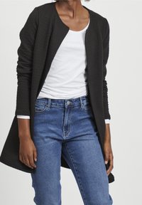 Svart texturerad lång cardigan med rund halsringning, kombinerad med en vit skjorta och blå denimjeans, med rak skärning och framfickor.