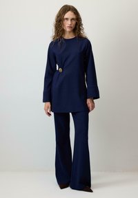 Tunika blu navy con maniche lunghe, una piega laterale e un accento circolare dorato. Abbinata a pantaloni a palazzo coordinati realizzati in tessuto morbido.