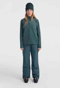Světlý fleece svetr s zipem a dlouhými rukávy, zkombinovaný s odpovídajícími světlými sněhovými kalhotami a černými lyžařskými botami, doplněný pletenou čepicí.