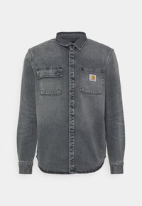Camisa de mezclilla gris con mangas largas, cuello con botones, dos bolsillos en el pecho y un pequeño parche con logo en el bolsillo izquierdo. Diseño clásico.