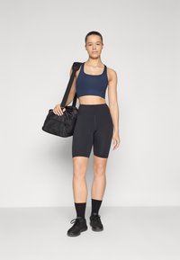 Idrettsantrekk bestående av en navy crop top og svarte sykkelshorts. Modellen holder en svart duffelbagg med teksturert overflate.