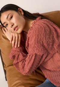 Blouse transparente couleur rouille avec des pois texturés, des poignets à volants et des détails en dentelle florale. Portée avec un denim clair, stylée de manière décontractée.