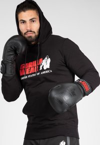 Haut à capuche noir avec le logo rouge "Gorilla Wear", associé à des gants de boxe noirs texturés. Le modèle pose avec confiance, les poings levés.