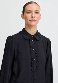 Blusa nera con colletto arricciato e patta frontale. Maniche lunghe con spalle arricciate. Tessuto leggero con una texture liscia.