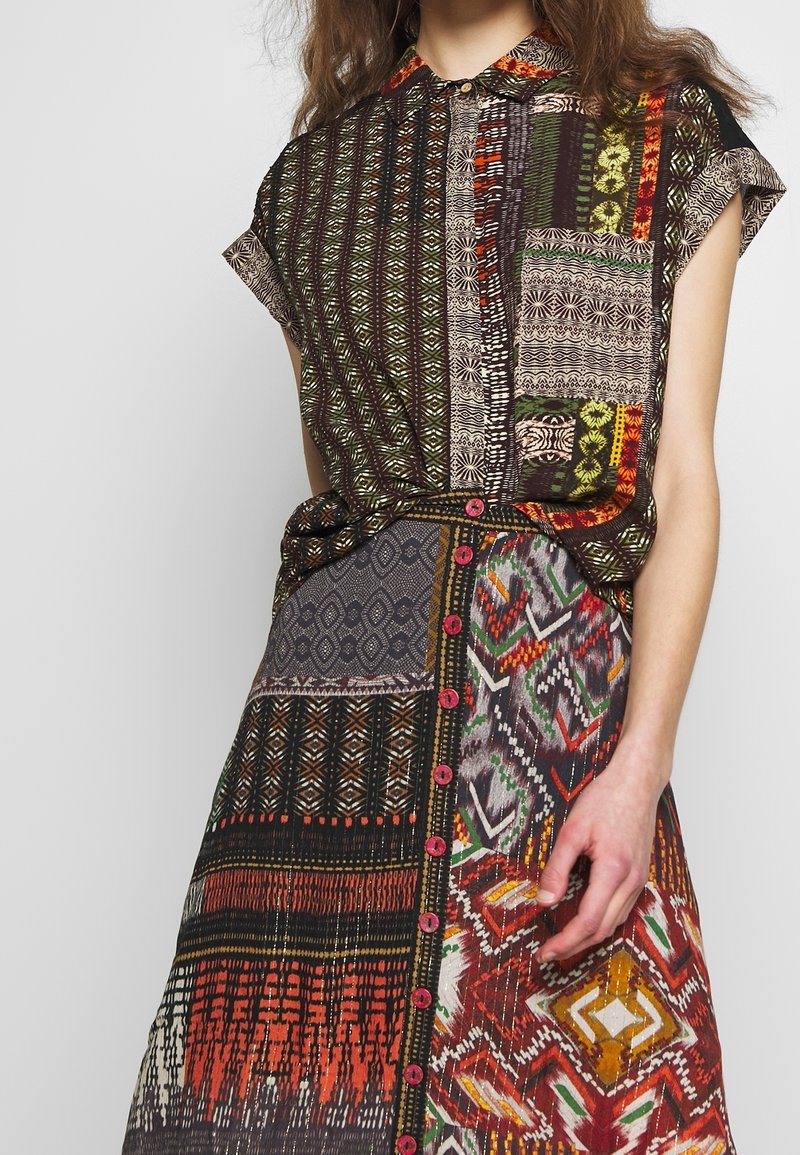 Femme portant une robe patchwork multicolore avec des motifs géométriques et tribaux, manches courtes, boutons sur le devant et une poche poitrine.