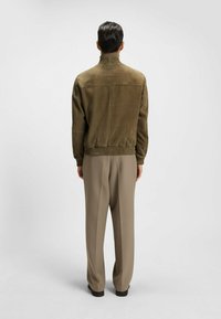 Olijfgroene suède jas met een hoge kraag en geribde manchetten, gecombineerd met beige wijde broekspijpen. Zachte textuur en ontspannen pasvorm.