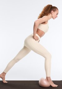 Frau in beige Sportbekleidung, die ein Bein nach vorne streckt und den Fuß auf einem pinken halbballförmigen Balance-Trainer auf einer dunklen Trainingsmatte abstützt.