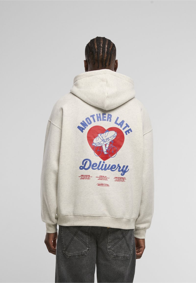 Personne avec des cheveux tressés portant un sweat à capuche clair avec un graphisme de cœur rouge, chaussure et bicyclette et le texte « Another Late Delivery » au dos.