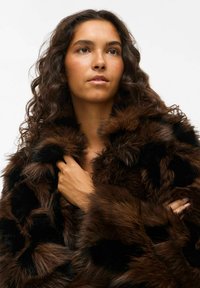 Brūnas un melnas mākslīgās kažokādas mēteli, kas izceļas ar teksturētu, apjomīgu dizainu. Mētelim ir lieli apkakles akcenti un grezna, ērtā izskats.