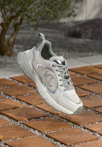 Hellgrauer Sportschuh mit Mesh- und synthetischen Überzügen, runder Zehenkappe, gepolstertem Kragen und weißer Sohle, präsentiert auf strukturierten braunen Fliesen.