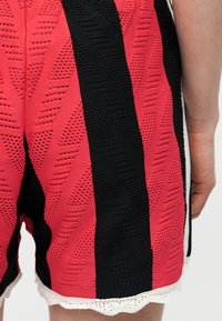 Κόκκινο και μαύρο πλεκτό σορτς με υφασμένους γεωμετρικούς σχεδιασμούς και λευκή διακοσμητική μπορντούρα σε κυματιστό σχέδιο, φορεμένο από ένα άτομο, με εμφανές το χέρι δίπλα στα σορτς.