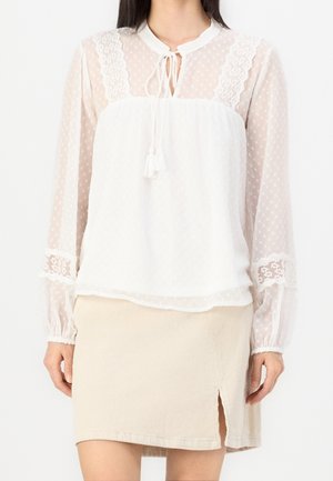 Femme portant un chemisier blanc transparent avec des détails en dentelle et une jupe en velours côtelé beige avec une fente devant, debout devant un fond uni.