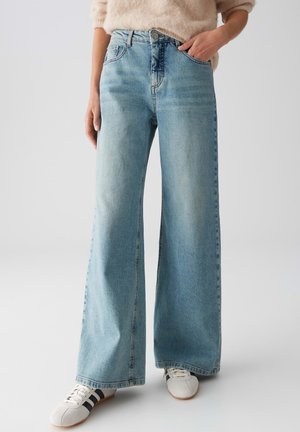 Lichtblauwe wijduitlopende denimjeans met een hoge taille, vijf zakken en subtiele vervaging. Gekleed met witte sneakers.
