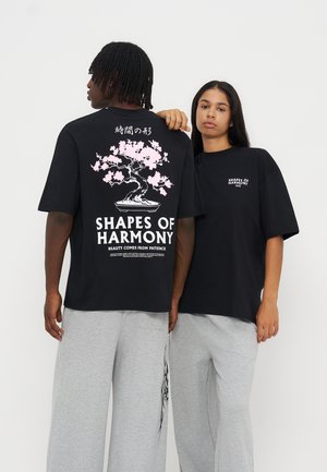 Deux modèles portent des t-shirts noirs oversize avec un bonsaï rose et le texte "Shapes of Harmony", associés à un pantalon large gris clair.