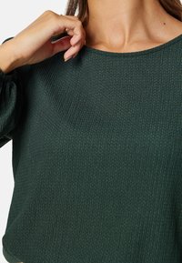 Ciemnozielony, prążkowany top z szerokim dekoltem i bufkami, wykonany z teksturowanej tkaniny. Zbliżenie na dekolt i część dłoni.