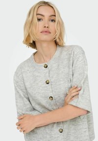 Femme blonde aux cheveux courts portant un pull gris clair à boutons, les bras croisés sur la poitrine, devant un fond neutre.